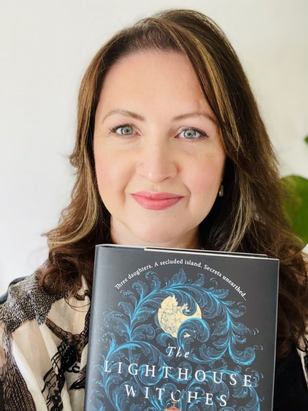 CJ Cooke faces the camera. She is a white woman with brown shoulder length hair and blue-green eyes, and is smiling. She holds a copy of her novel THE LIGHTHOUSE WITCHES.