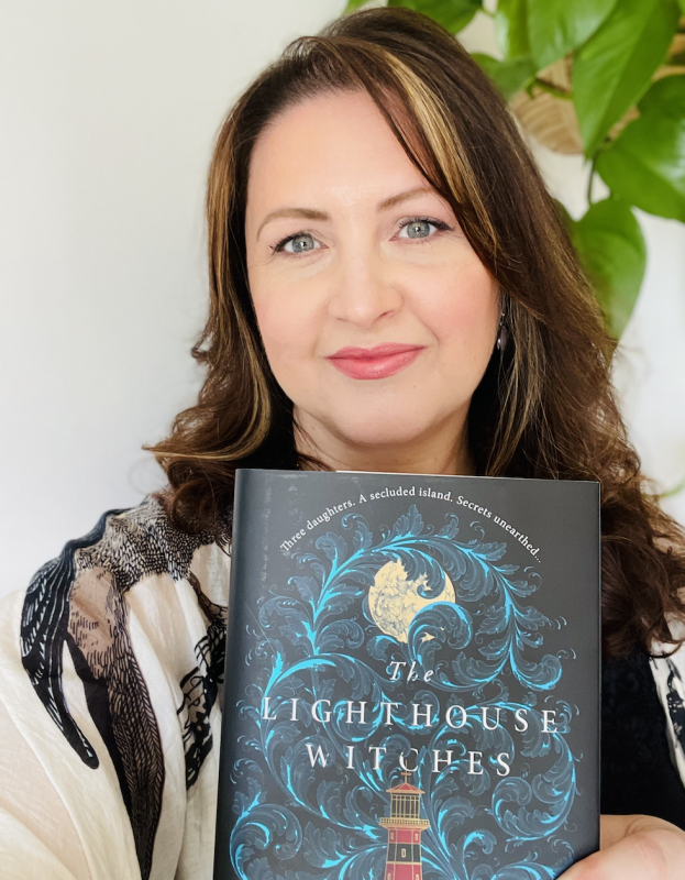 CJ Cooke, a white woman with dark brown hair, blue eyes and pink lipstick, smiles at the camera while holding a copy of The Lighthouse Witches